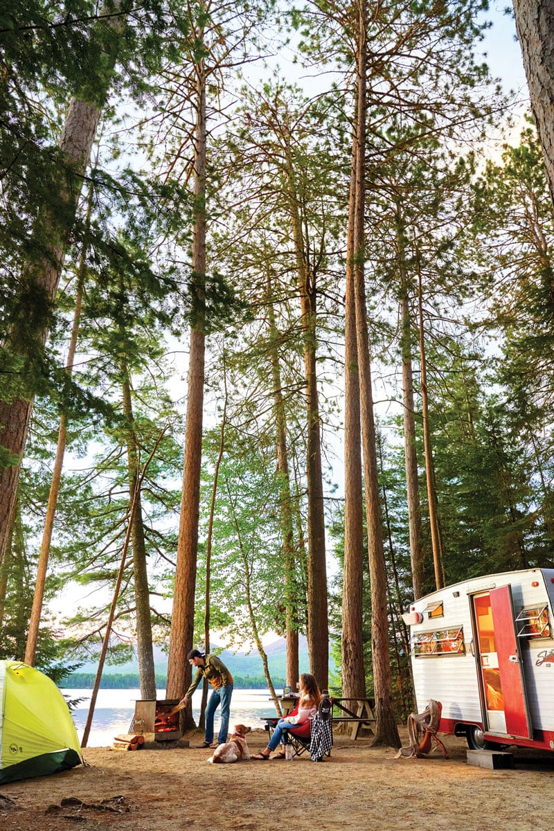 A group of people sitting around a camper in the woods in Cathedral Pines Campground.