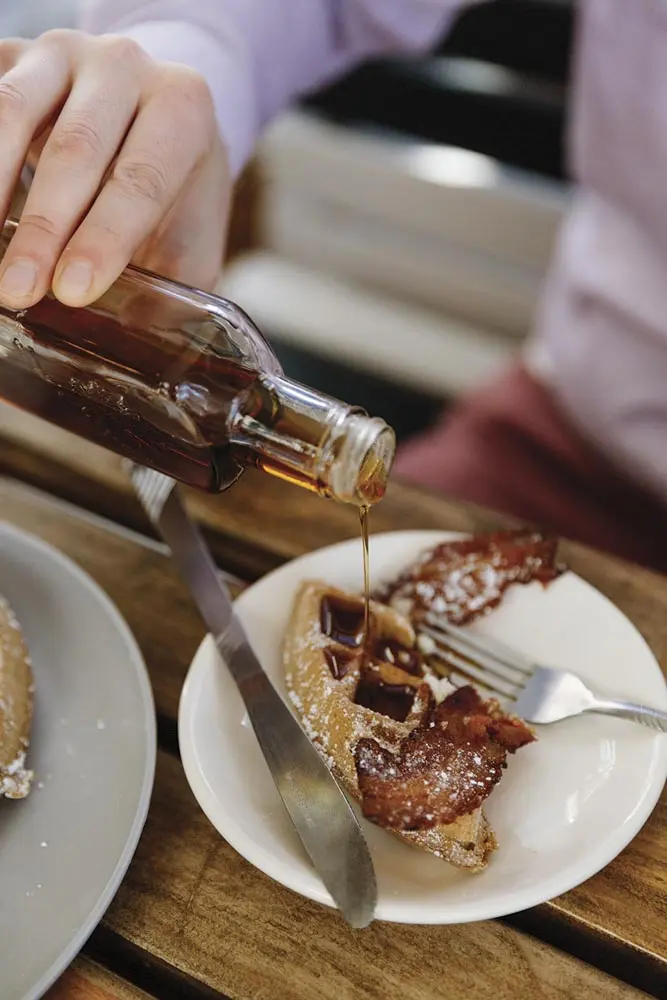 syrup being poured onto a waffle section topped with bacon