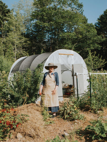 Amy Pollien stands in front of a hoop house where she keeps art supplies so she can set up her easel in the garden. At her feet is a mound of composting garden waste and other organic materials — a future vegetable bed.