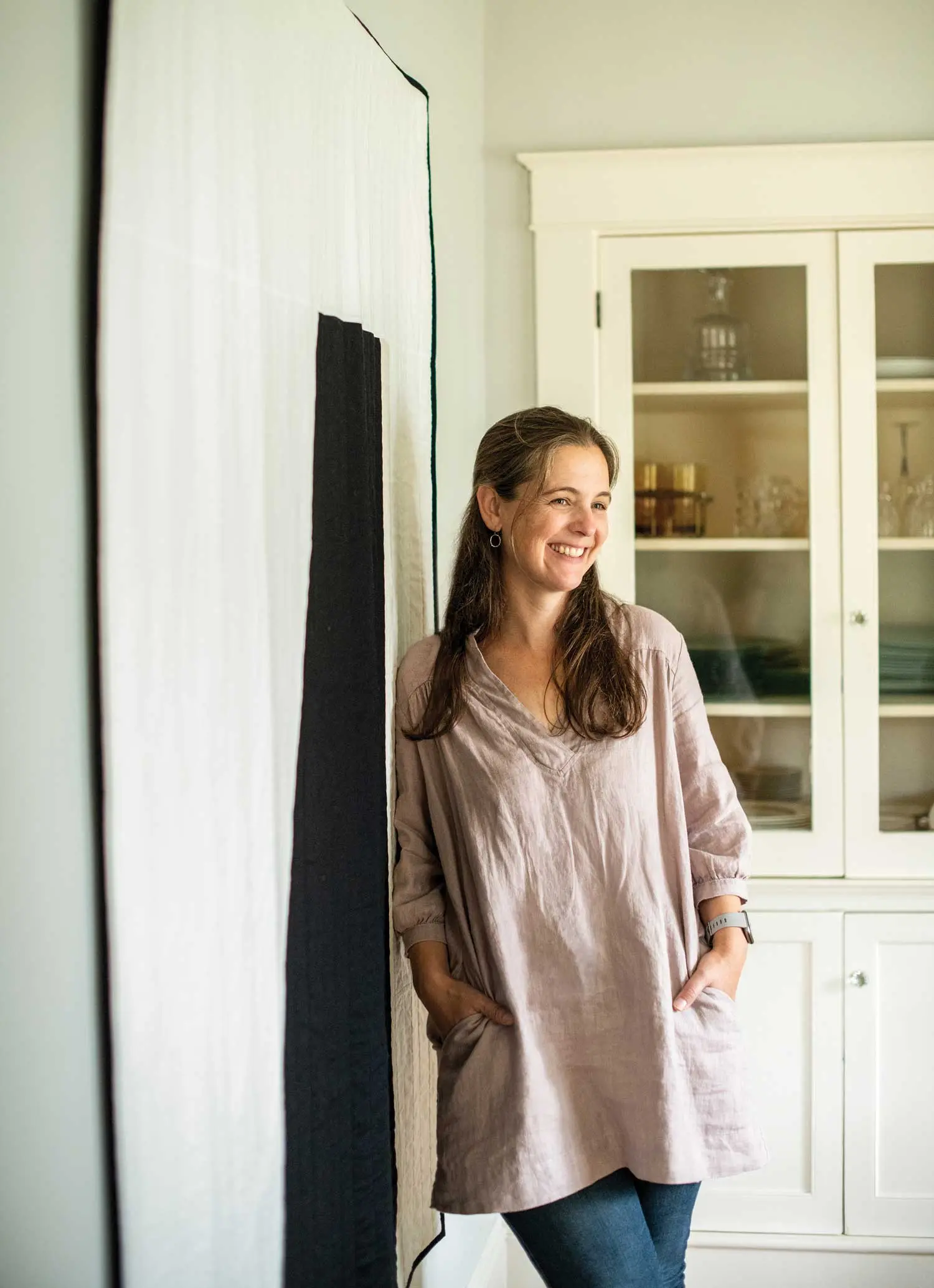 Dash Masland, owner of Smith's General, leans against the Mackworth quilt “mural” in her Yarmouth dining room; linen curtains inspired by ancient Korean patchworks hang nearby
