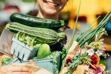 a woman in sunglasses holding all of her summer produce purchases from a Maine farmers market