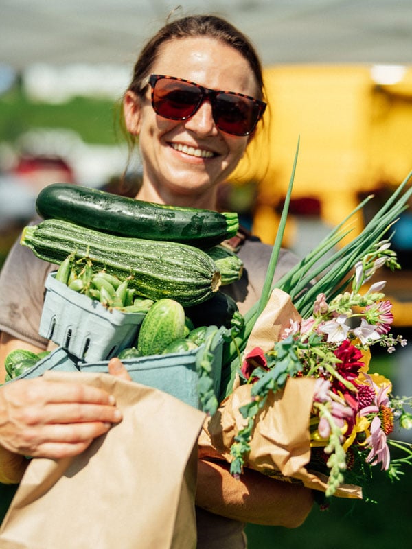 a woman in sunglasses holding all of her summer produce purchases from a Maine farmers market