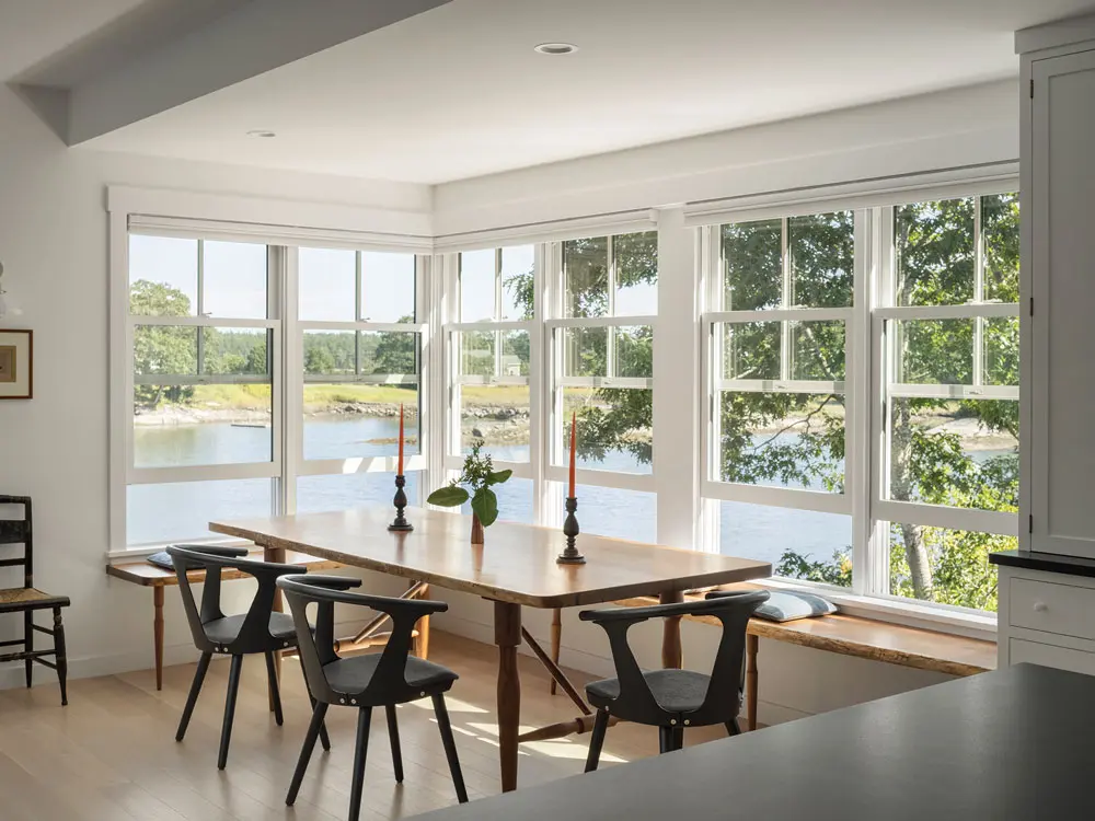 In the dining room, Wraparound windows turn Frost Fish Cove into a mural in the dining space, furnished with a reclaimed-walnut table and L-shaped bench by Biddeford woodworker Derek Preble.
